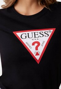 Černé bavlněné tričko s velkým trojúhelníkovým bílým logem s červenými akcenty, na kterém je nápis "GUESS U.S.A." a červený otazník.