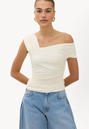 Frau trägt ein weißes, einärmeliges, gerafftes Oberteil und blaue, hoch taillierte Jeans mit goldenen Creolen, steht mit den Händen hinter dem Rücken.