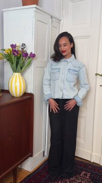 Mujer con cabello oscuro que lleva una chaqueta vaquera clara y pantalones negros, de pie en el interior cerca de un armario blanco y un jarrón a rayas amarillas con flores.