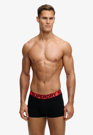 Muskelöser Mann steht, trägt schwarze Boxershorts mit rotem Bund und der Aufschrift "SUPERDRY" auf weißem Hintergrund.