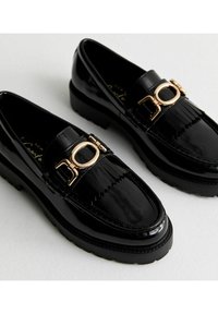 Mocasines de charol negro con una hebilla decorativa en tono dorado, que cuentan con un detalle de flecos texturizados y una suela de goma negra gruesa.