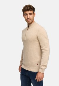 Beige stickad tröja med halv blixtlås, ribbade manschetter och nederkant, mjuk textur, med en liten läderlogga nära botten.