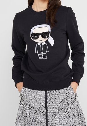 Žena v černé mikině s karikaturou mužské postavy v černém obleku, se světlými vlasy a slunečními brýlemi, doplněnou černo-bílou vzorovanou sukní.