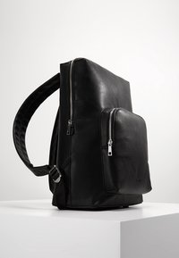 Svart läderryggsäck med en strukturerad form, med en framficka med dragkedja och texturerade axelremmar. Silverfärgade detaljer ingår.