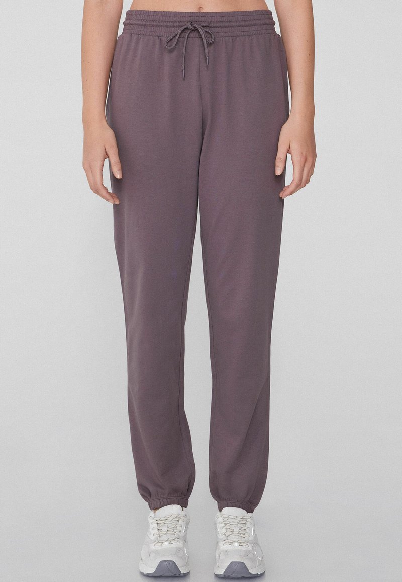 Pantalones deportivos morados con un acabado texturizado, que cuentan con una cintura elástica y dobladillos ajustados. Combinados con zapatillas deportivas gris claro.