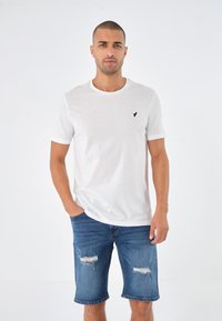White cotton t-shirt with a crew neck and black logo on the left chest, paired with distressed denim shorts in medium blue.