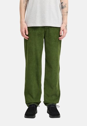 Personne portant un pantalon large en velours côtelé vert foncé, un t-shirt gris clair et des chaussures noires à lacets, avec des tatouages visibles sur les deux avant-bras.