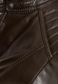 Tumepruun nahk materjal, millel on tekstureeritud pind, esile tõstetud õmblused, tikandiga aktsendid ja metallist nupuga detailid.