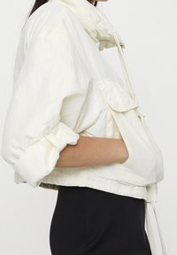 Krótka kurtka w kolorze kremowym z wysokim kołnierzem, ściągaczem oraz dużą przednią kieszenią. Charakteryzuje się podwiniętymi rękawami i luźnym krojem.