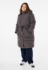 Padded gray coat with a belted waist, hood, and front pockets; features a button closure and quilted texture. Paired with dark jeans and sneakers.