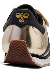 Schwarzer und metallisch goldener Sneaker mit glatter Textur. Hat ein Logo an der Ferse und eine braune Gummisohle mit Zickzackmuster.