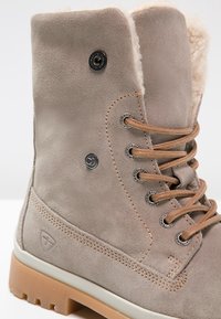 Jasnobrązowy zamszowy botek za kostkę z podszewką z sztucznego futra, okrągłymi metalowymi przelotkami i beżowymi sznurowadłami; posiada teksturowaną gumową podeszwę zapewniającą przyczepność.