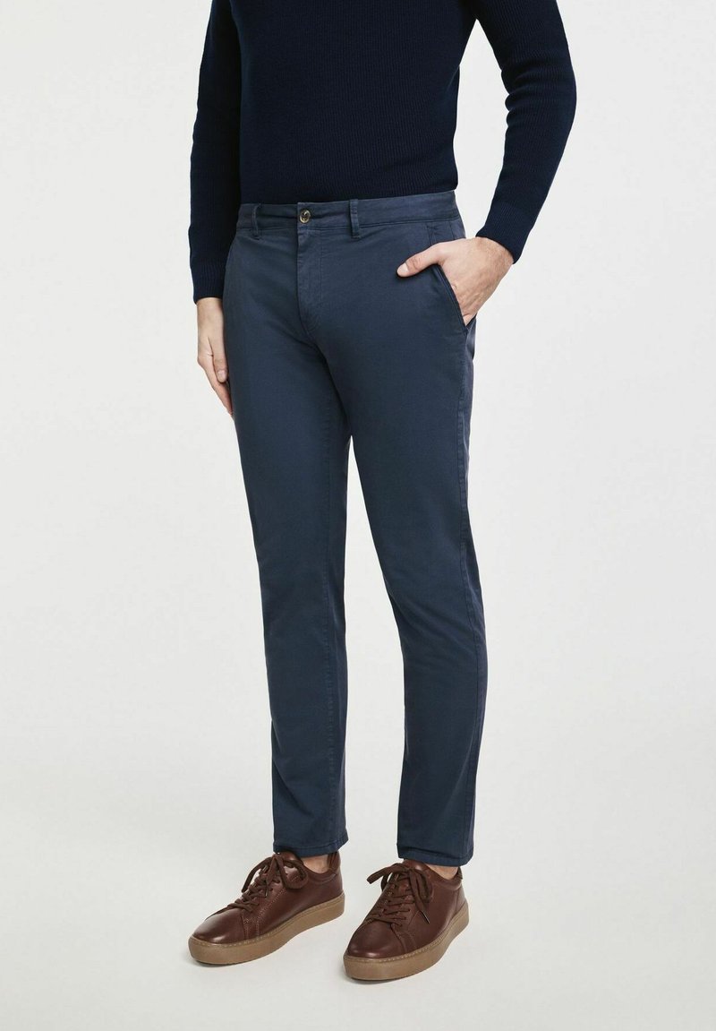 Mężczyzna stoi w ciemnym swetrze i granatowych chinosach, jedna ręka w kieszeni, w brązowych sneakersach na neutralnym tle.