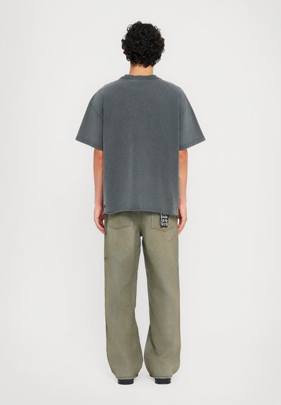 Pelēka oversized t-kreklu ar īsām piedurknēm, brīvs griezums, parādīts kopā ar vaļīgām olīvzaļu biksēm ar dekoratīvu plāksteri aizmugurē.