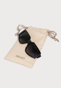 Gafas de sol negras de gran tamaño con lentes degradados, acompañadas de una bolsa de drawstring beige con el logotipo dorado "LIU · JO".