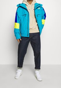 Kolorowa niebieska kurtka z żółtymi i niebieskimi akcentami, kieszeniami na zamek oraz regulowanymi mankietami. Połączona z beżową bluzą z kapturem i ciemnymi dżinsami.