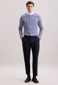Uomo in piedi con le mani in tasca, che indossa una camicia a righe blu e bianche, pantaloni blu scuro e scarpe eleganti nere.
