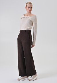 Lunghe pantaloni marroni con una superficie liscia, abbinati a un maglione beige scollato sulle spalle. Sneakers bianche con accenti sottili.