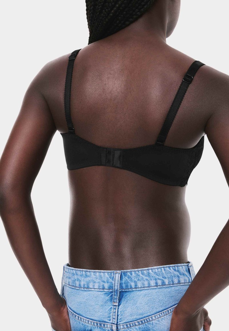 Sort blonde-bh med justerbare stropper og lukning med hægte. Modellen har lyseblå denimjeans på. Glat stof med teksturerede detaljer.