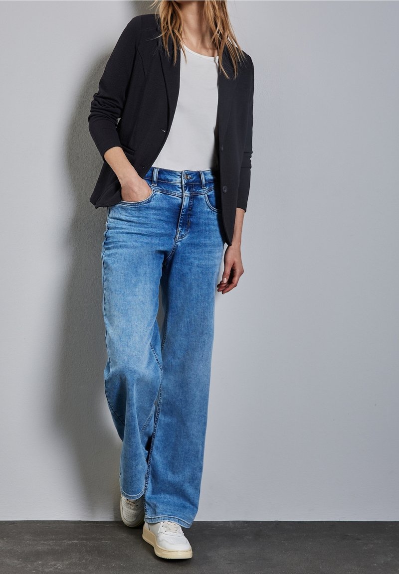 Femme portant un blazer noir, une chemise blanche, un jean bleu à jambes larges et des baskets blanches, debout contre un mur gris uni.