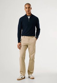 Temno modra majica z zadrgo, bež hlače in bele superge s črnimi poudarki. Gladek material, oprijet kroj in subtilen detajl na manšetah so vidni.