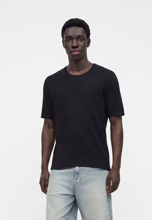 Jeune homme aux cheveux courts et bouclés, portant une chemise noire à manches courtes et un jean bleu clair sur un fond uni.