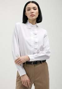 Blusa bianca a maniche lunghe con colletto arricciato e chiusura a bottoni, abbinata a pantaloni a righe marrone chiaro e una cintura nera. Texture del tessuto liscia.