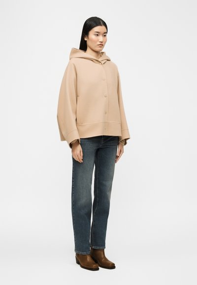 Γυναίκα που στέκεται φορώντας μπεζ μπουφάν με κουκούλα και κουμπιά, μπλε τζιν και καφέ μποτάκια αστραγάλου πάνω σε απλό λευκό φόντο.