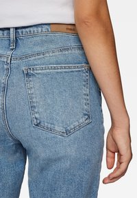 Los vaqueros de mezclilla azul claro tienen un corte recto, un bolsillo trasero izquierdo con solapa puntiaguda y una etiqueta de cuero marrón con la marca en la cintura.