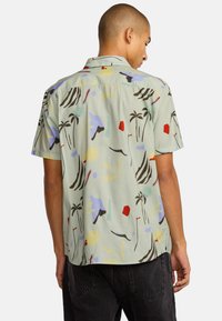 Korte mouw shirt in lichtgroene stof met een kleurrijk, abstract patroon van palmbomen, geometrische vormen en zonmotieven.