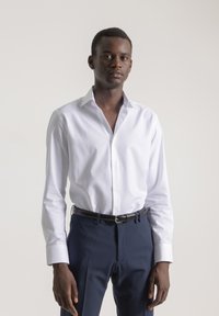 Joven de pie, vistiendo una camisa blanca de botón metida dentro de pantalones azul marino con un cinturón negro, frente a un fondo claro y liso.