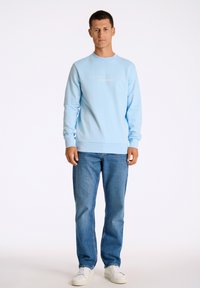 Ljusblå sweatshirt med rund halsringning och logotyp, tillsammans med ljusblå jeans och vita sneakers. Slät textur, avslappnad passform.