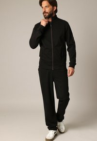 Zwarte ritsjack met hoge kraag, gecombineerd met bijpassende zwarte broek. Het jack heeft een contrasterende rits en zijzak. Witte sneakers.