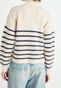 Suéter de punto en color crema con rayas horizontales azul marino; cuenta con un cuello alto y un corte holgado. Combinado con jeans de mezclilla azul claro.