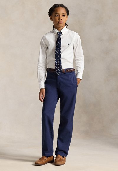 Garçon portant une chemise blanche, une cravate bleu marine avec motif de bouclier, un pantalon bleu marine, une ceinture tressée marron et des mocassins marron, debout avec une main dans la poche.