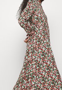 Donna che indossa un vestito a maniche lunghe con colletto, caratterizzato da un motivo floreale rosso, bianco e verde, in piedi con le mani dietro la schiena.