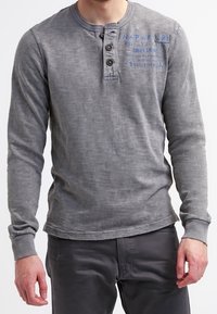 Mann in einem verblassten grauen Langarm-Henley-Shirt mit drei Knöpfen und blauer Schrift auf der Brust, kombiniert mit dunklen Hosen.