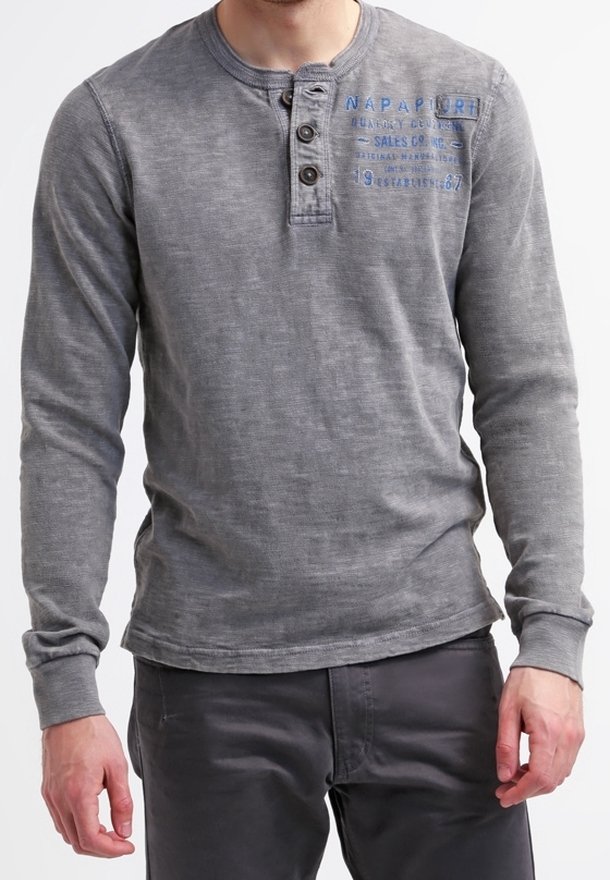 Mann in einem verblassten grauen Langarm-Henley-Shirt mit drei Knöpfen und blauer Schrift auf der Brust, kombiniert mit dunklen Hosen.