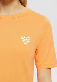 Orange t-shirt i bomullstyg, med rund halsringning och korta ärmar. Tryckt vit text i form av ett hjärta som säger "ALLT VI BEHÖVER ÄR NAPPA."