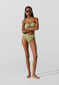 Olivzaļš divdaļīgs bikini ar rušētu bandeau topu ar centrālu akcentu un augsta vidukļa apakšdaļu, izceļot spīdīgu tekstūru.