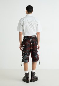 Camisa de manga corta a rayas, pantalones cortos cargo de camuflaje negro y rojo, torso visible desde la parte de atrás y botas negras con detalles en blanco.