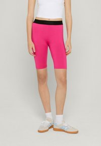 Světlé růžové cyklistické šortky s černým elastickým pasem označeným "SWIM SENSE", nošeny s bílým cropped topem a pohodlnými teniskami.