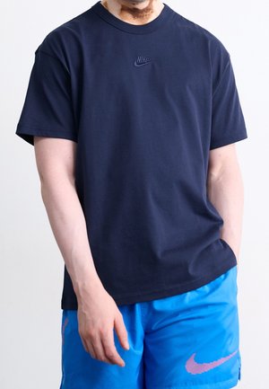 Mand iført en marineblå Nike t-shirt og lyseblå Nike shorts med et pink swoosh-logo på en hvid baggrund.
