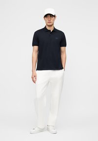 Młody mężczyzna ubrany w czarną koszulkę polo, białe spodnie, białe tenisówki i białą czapkę, stojący na jednolitym jasnym tle.