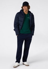 Chaqueta de abrigo azul marino, sudadera verde oscura, pantalones de chándal azul marino y zapatillas blancas. Incluye capucha y logo bordado en la ropa.