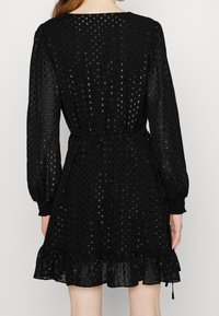 Svart, halvgenomskinlig klänning med texturerade prickar, långa puffärmar, åtsittande midja och volangslut. Har rund halsringning och justerbara knytband.