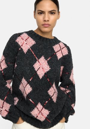 Kobieta w ciemnoszarym swetrze z dużymi różowymi wzorami w kształcie diamentu i czerwonymi cekinowymi akcentami, ręce w kieszeniach, neutralne tło.