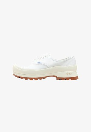 Bílé plátěné tenisky s tlustou, texturovanou gumovou mezipodešví a hnědou podrážkou s protiskluzovým vzorkem; mají klasický design na šněrování a detail loga.