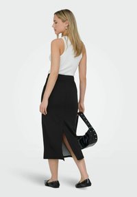 Schwarzer Rock mit hohem Taillenbund und einem seitlichen Schlitz, aus glattem Stoff. Kombiniert mit einem eng anliegenden weißen Tanktop und schwarzen Ballerinas, mit einer schwarzen Tasche.