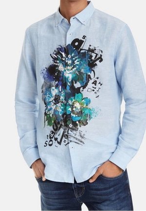 Camisa de botones azul claro con diseño floral azul y verde y texto abstracto negro en la parte delantera, combinada con vaqueros azul oscuro.